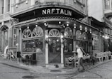 A cozy, corner café with a vintage exterior featuring arched windows and a string of lights along the awning. The scene includes outdoor seating with various chairs and tables, a passerby walking by, and a cat lying on the ground near the entrance. The setting suggests an urban environment with cobblestone streets.