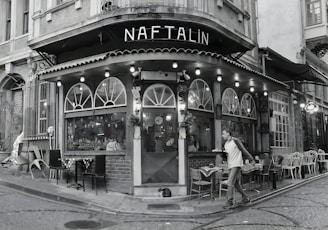 A cozy, corner café with a vintage exterior featuring arched windows and a string of lights along the awning. The scene includes outdoor seating with various chairs and tables, a passerby walking by, and a cat lying on the ground near the entrance. The setting suggests an urban environment with cobblestone streets.