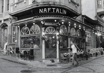 A cozy, corner café with a vintage exterior featuring arched windows and a string of lights along the awning. The scene includes outdoor seating with various chairs and tables, a passerby walking by, and a cat lying on the ground near the entrance. The setting suggests an urban environment with cobblestone streets.