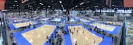 A large indoor sports facility features numerous volleyball courts arranged in a grid pattern. The courts are surrounded by spectators and players, with blue flooring and wooden court surfaces. Overhead lights illuminate the expansive space, and the location is bustling with activity.