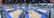 A large indoor sports facility features numerous volleyball courts arranged in a grid pattern. The courts are surrounded by spectators and players, with blue flooring and wooden court surfaces. Overhead lights illuminate the expansive space, and the location is bustling with activity.