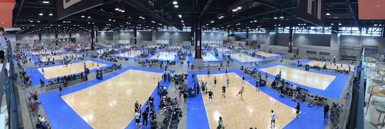 A large indoor sports facility features numerous volleyball courts arranged in a grid pattern. The courts are surrounded by spectators and players, with blue flooring and wooden court surfaces. Overhead lights illuminate the expansive space, and the location is bustling with activity.