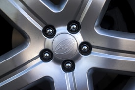 A close-up view of a car alloy wheel featuring a five-spoke design with a central emblem. The wheel has a metallic finish, and there are visible lug nuts securing it.