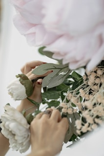 Hands carefully arranging a bouquet of delicate pink and white flowers, surrounded by green leaves and smaller beige blossoms.