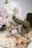 A person with curly hair is arranging large, pastel-colored paper flowers. The flowers include shades of white, pink, and beige, and they appear to be part of a decorative display. The scene has a soft and delicate feel, capturing a creative process.