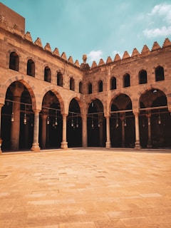 A historical building with impressive arched architecture surrounding an open courtyard. The arches are supported by slender columns, and the stone structure features intricate designs characteristic of ancient craftsmanship. The sky is a vibrant blue with some scattered clouds, adding contrast to the earthy tones of the building.