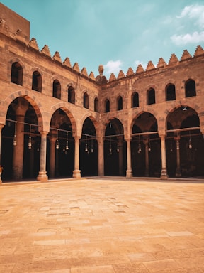 A historical building with impressive arched architecture surrounding an open courtyard. The arches are supported by slender columns, and the stone structure features intricate designs characteristic of ancient craftsmanship. The sky is a vibrant blue with some scattered clouds, adding contrast to the earthy tones of the building.