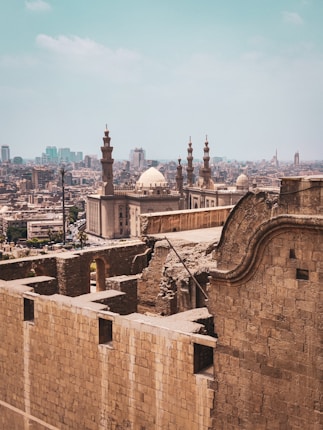 Ancient stone walls and structures overlook a sprawling cityscape with a prominent mosque featuring minarets and a dome visible in the foreground. The city extends into the distance with modern buildings against a clear blue sky.