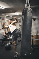A person is engaging in a kickboxing workout with a heavy punching bag. He is raising his leg to deliver a kick while wearing a sleeveless shirt and shorts in a gym setting. Another individual is visible in the background, seated on a bench, seemingly resting between exercises.
