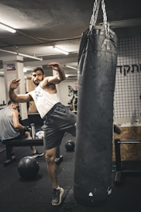 A person is engaging in a kickboxing workout with a heavy punching bag. He is raising his leg to deliver a kick while wearing a sleeveless shirt and shorts in a gym setting. Another individual is visible in the background, seated on a bench, seemingly resting between exercises.