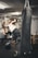 A person is engaging in a kickboxing workout with a heavy punching bag. He is raising his leg to deliver a kick while wearing a sleeveless shirt and shorts in a gym setting. Another individual is visible in the background, seated on a bench, seemingly resting between exercises.