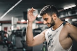 A man with a muscular build is wearing a sleeveless shirt and appears to be in a gym setting. He is smiling slightly with his left arm raised. Exercise equipment is visible in the background, suggesting an indoor workout environment.