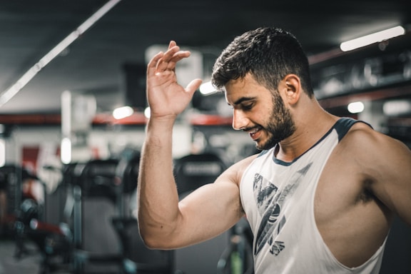 A man with a muscular build is wearing a sleeveless shirt and appears to be in a gym setting. He is smiling slightly with his left arm raised. Exercise equipment is visible in the background, suggesting an indoor workout environment.