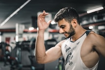 A man with a muscular build is wearing a sleeveless shirt and appears to be in a gym setting. He is smiling slightly with his left arm raised. Exercise equipment is visible in the background, suggesting an indoor workout environment.