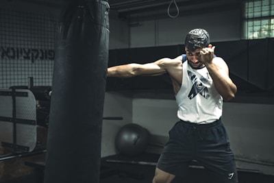A man in a gym is intensely training with a punching bag. He appears focused and determined, wearing a sleeveless top and shorts. The setting is an indoor gym with exercise equipment in the background.