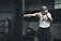 A man in a gym is intensely training with a punching bag. He appears focused and determined, wearing a sleeveless top and shorts. The setting is an indoor gym with exercise equipment in the background.