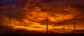Electricity pylons and wires stretch across a scenic landscape during sunset, with bold, fiery colors lighting up the dramatic clouds in the sky. The silhouette of distant mountains provides a striking backdrop.