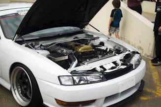 A car with steam rising from the hood parked on a roadside, with a concerned man standing nearby scratching his head.