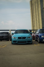 A turquoise car is prominently parked in an outdoor parking lot, flanked by other cars including a dark gray car on the left and a blue car on the right. Part of a tall building can be seen in the background, under a partially cloudy sky.