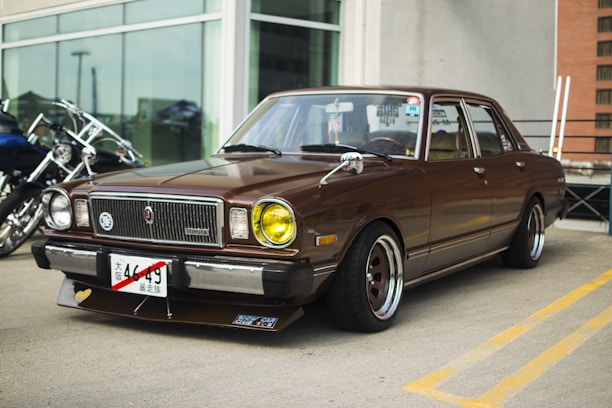 A vintage brown car is parked outdoors, featuring a boxy design with distinctive yellow headlights and aftermarket wheels. The front license plate has Japanese characters, and there are reflections on the car's windshield. The background shows part of a modern building and some motorcycles parked nearby.