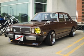 A vintage brown car is parked outdoors, featuring a boxy design with distinctive yellow headlights and aftermarket wheels. The front license plate has Japanese characters, and there are reflections on the car's windshield. The background shows part of a modern building and some motorcycles parked nearby.