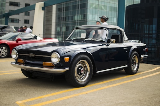 A classic convertible car with a dark blue exterior is parked in an urban setting. The car features chrome accents and retro design elements, with its top down. In the background, a modern glass building is visible along with a red sports car and a few people standing nearby.