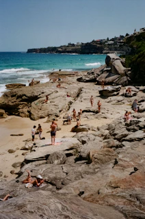 A rocky beach scene with people sunbathing and enjoying the coastal environment. The ocean is visible in the background, with waves gently breaking near the shore. The coastline stretches into the distance, bordered by cliffs and scattered trees and buildings.