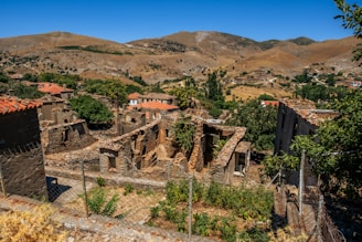 An old, abandoned village with stone ruins and partially collapsed buildings is nestled in a rugged, mountainous landscape. The structures are surrounded by sparse vegetation, with some greenery and trees scattered among them. In the background, hills and distant mountains create a stark contrast with the blue sky.