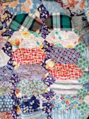 A patchwork quilt featuring an assortment of colorful, patterned fabric pieces. The patterns include floral designs, checkered stripes, and polka dots in various shades including blues, reds, oranges, and greens. The quilt appears to be handmade with intricate stitching connecting the different swatches.