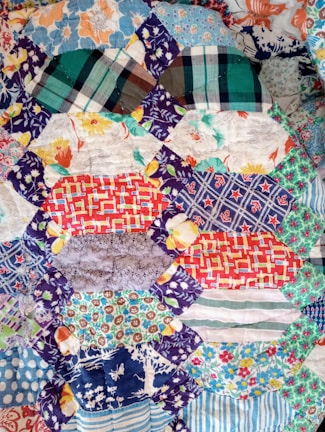 A patchwork quilt featuring an assortment of colorful, patterned fabric pieces. The patterns include floral designs, checkered stripes, and polka dots in various shades including blues, reds, oranges, and greens. The quilt appears to be handmade with intricate stitching connecting the different swatches.