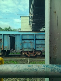 A blue freight train car is parked on railway tracks beneath a metal bridge. The background includes greenery and a clear sky, while the foreground shows a rusted metal railing.