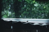 Raindrops fall heavily, creating a blurred effect against a background of green foliage. The image captures the view from beneath a corrugated roof, with water bouncing off the surface and streaming down.