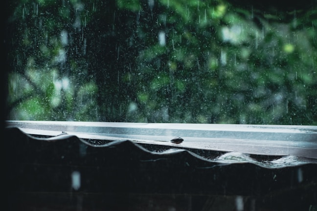 Raindrops fall heavily, creating a blurred effect against a background of green foliage. The image captures the view from beneath a corrugated roof, with water bouncing off the surface and streaming down.