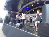 A band performs on an outdoor stage under a canopy, featuring multiple musicians with various instruments including a saxophone, trumpet, and guitar. The stage is equipped with large speakers and microphones, set against a backdrop with partial text. The musicians appear casually dressed and one female musician stands to the right, adjusting her sunglasses.