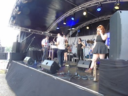 A band performs on an outdoor stage under a canopy, featuring multiple musicians with various instruments including a saxophone, trumpet, and guitar. The stage is equipped with large speakers and microphones, set against a backdrop with partial text. The musicians appear casually dressed and one female musician stands to the right, adjusting her sunglasses.