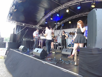 A band performs on an outdoor stage under a canopy, featuring multiple musicians with various instruments including a saxophone, trumpet, and guitar. The stage is equipped with large speakers and microphones, set against a backdrop with partial text. The musicians appear casually dressed and one female musician stands to the right, adjusting her sunglasses.