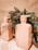 Two peach-colored shampoo bottles are placed on a marble surface. The bottles are labeled 'Monday Moisture Shampoo' and are surrounded by green leafy plants, adding a natural and fresh contrast to the scene.