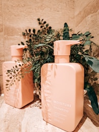 Two peach-colored shampoo bottles are placed on a marble surface. The bottles are labeled 'Monday Moisture Shampoo' and are surrounded by green leafy plants, adding a natural and fresh contrast to the scene.