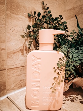 A peach-colored bottle of Monday Conditioner is placed on a stone tile surface, surrounded by green leafy plants. The bottle's design is minimalistic with embossed branding and text indicating it is PETA certified and contains 350ml. The natural background gives a calming and organic feel to the setting.