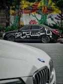 A black car with Arabic script painted on its side is parked on the street. Behind it, a vibrant mural with abstract and floral patterns decorates a wall. In the foreground, part of a white car with a BMW logo is visible.