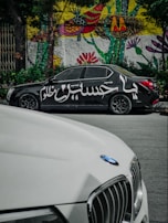 A black car with Arabic script painted on its side is parked on the street. Behind it, a vibrant mural with abstract and floral patterns decorates a wall. In the foreground, part of a white car with a BMW logo is visible.