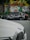 A black car with Arabic script painted on its side is parked on the street. Behind it, a vibrant mural with abstract and floral patterns decorates a wall. In the foreground, part of a white car with a BMW logo is visible.