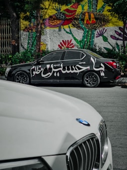 A black car with Arabic script painted on its side is parked on the street. Behind it, a vibrant mural with abstract and floral patterns decorates a wall. In the foreground, part of a white car with a BMW logo is visible.