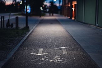 A dimly lit urban pathway with painted arrows indicating direction. The path is bordered by a building on the right and a few street poles on the left. In the background, city lights and blurred outlines of trees and other urban elements can be seen, typical of an evening or night scene.