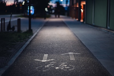 A dimly lit urban pathway with painted arrows indicating direction. The path is bordered by a building on the right and a few street poles on the left. In the background, city lights and blurred outlines of trees and other urban elements can be seen, typical of an evening or night scene.