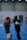 Two people are engaged in a boxing sparring session in an indoor sports facility. One person is wearing red gloves and the other is wearing white gloves and a protective headgear. The person with red gloves appears to be on the defensive, while the other is in an attacking position. The background reveals a blue flooring typical of indoor sports courts, with other individuals visible further away.