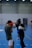 Two people are engaged in a boxing sparring session in an indoor sports facility. One person is wearing red gloves and the other is wearing white gloves and a protective headgear. The person with red gloves appears to be on the defensive, while the other is in an attacking position. The background reveals a blue flooring typical of indoor sports courts, with other individuals visible further away.