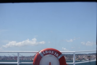 A red life buoy labeled 'AROEIRA' is mounted on the railing of a boat, with a view of a harbor and a city skyline in the background under a clear blue sky.