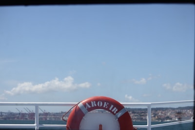 A red life buoy labeled 'AROEIRA' is mounted on the railing of a boat, with a view of a harbor and a city skyline in the background under a clear blue sky.