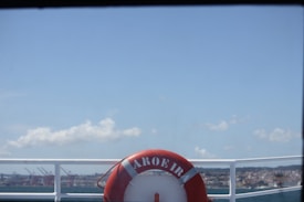 A red life buoy labeled 'AROEIRA' is mounted on the railing of a boat, with a view of a harbor and a city skyline in the background under a clear blue sky.